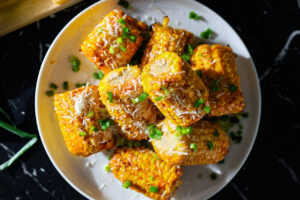 Grillissä kullanruskeiksi grillatut maissit parmesaanijuustolla ja keväsipulilla koristeltuna valkoisella lautasella.